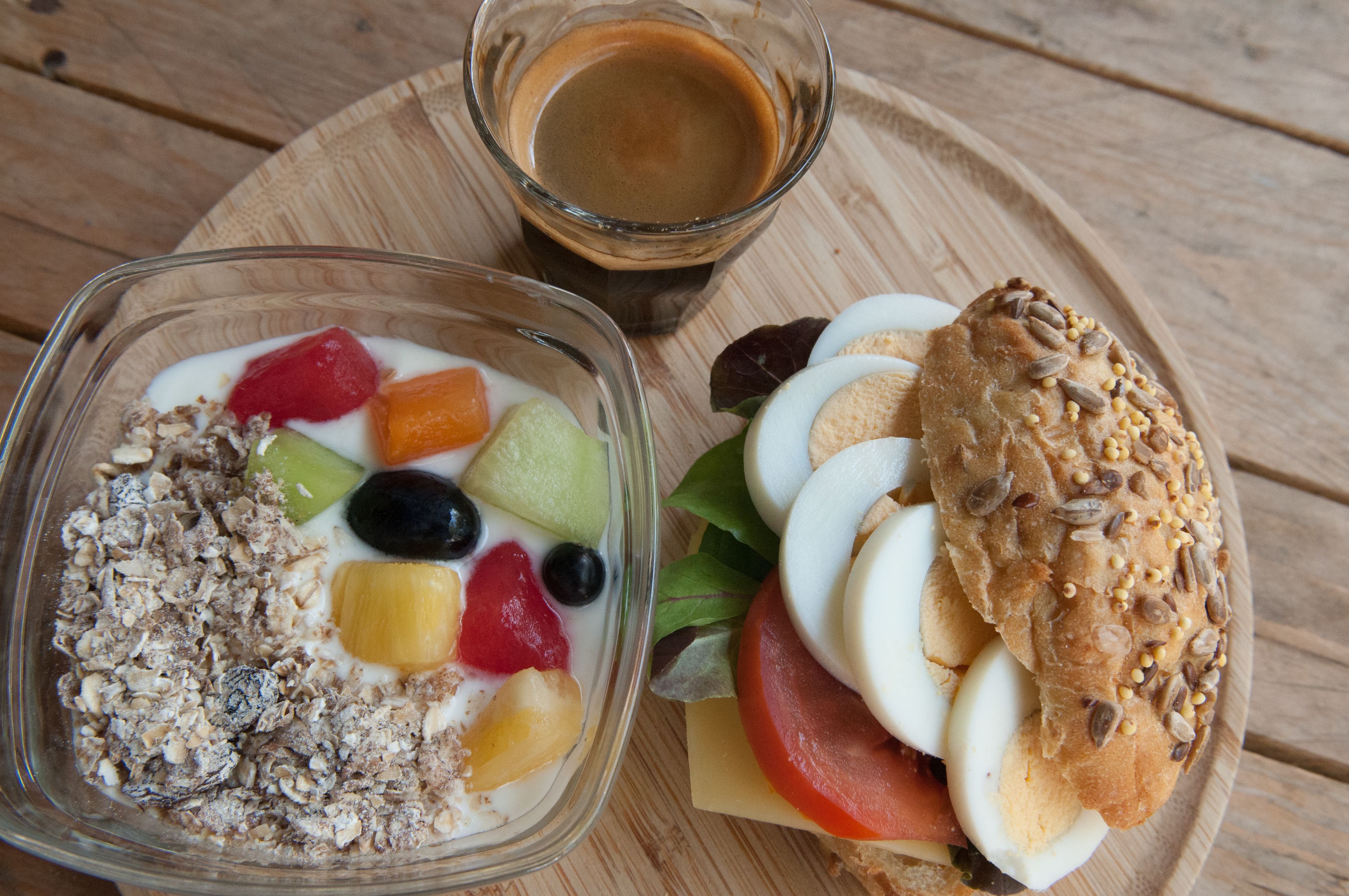Una comida que incluye una ensalada de frutas, un sándwich y una taza de café sobre una mesa de madera.