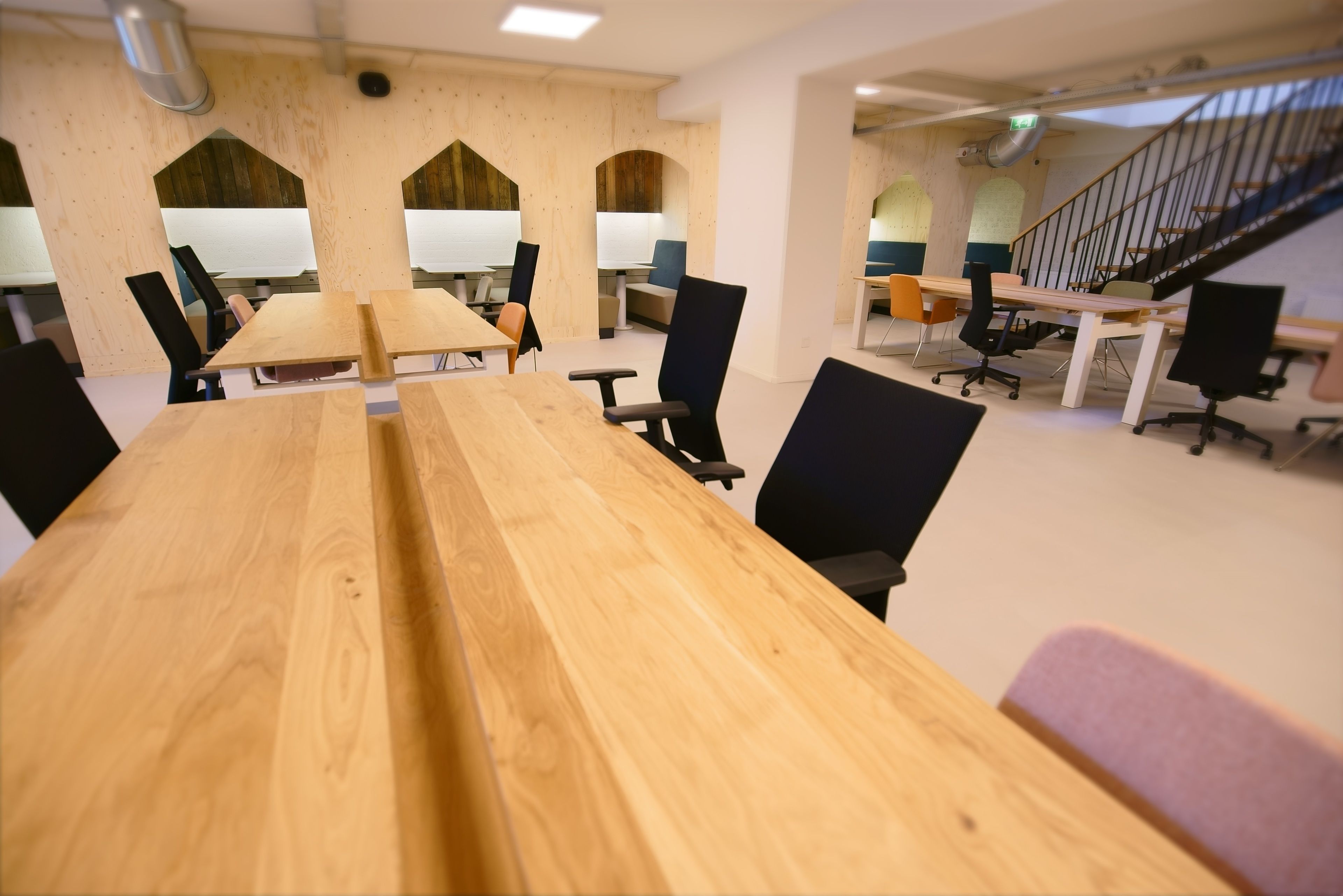 A modern conference room with wooden tables and black chairs, featuring a staircase and arched windows in the background.