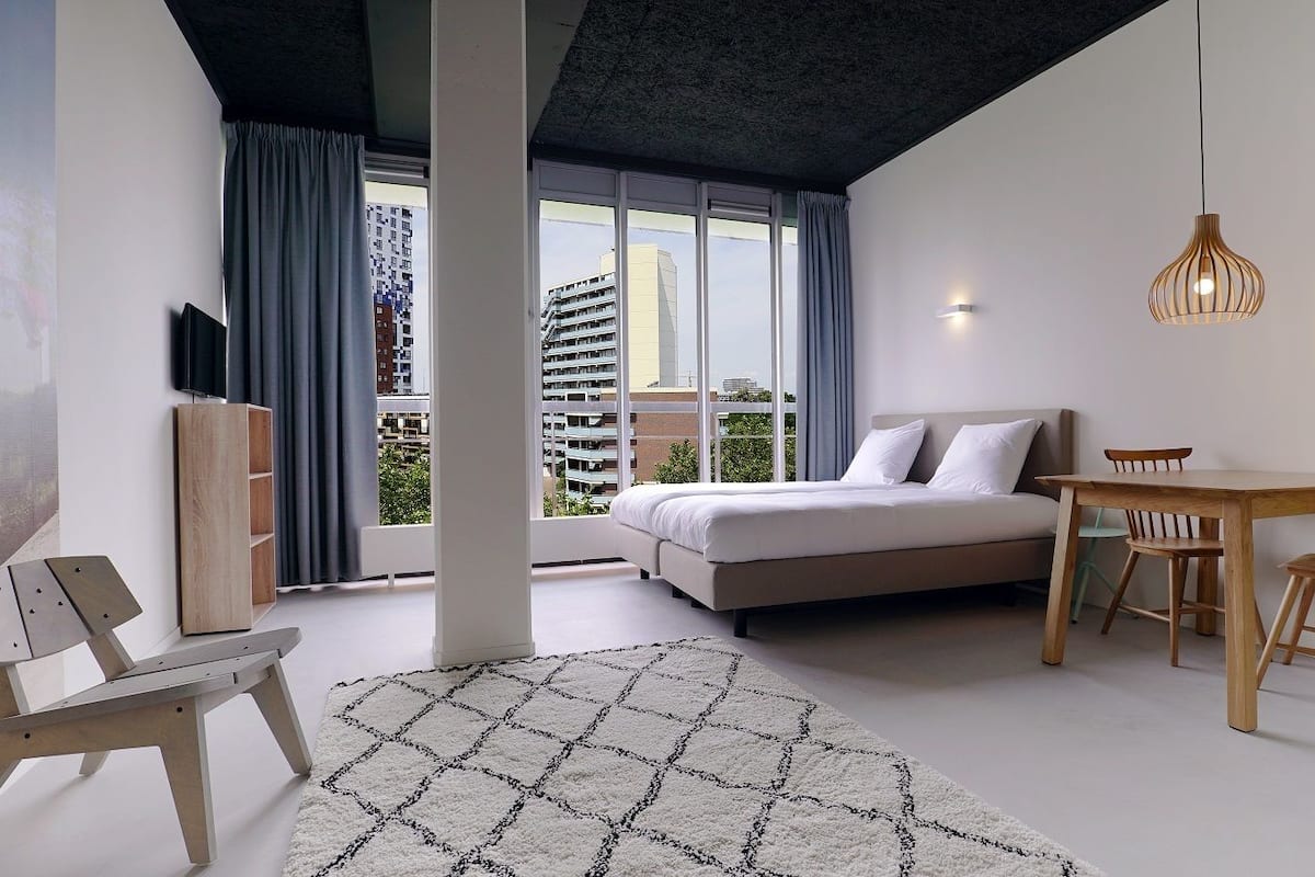 Een moderne slaapkamer met een bed, een eettafel en een stoel. Er is een groot raam met gordijnen, een plafondlamp en een aan de muur bevestigde televisie.