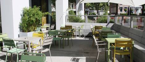 An outdoor seating area with green and yellow chairs and tables, a large umbrella, and a view of buildings and trees in the background.