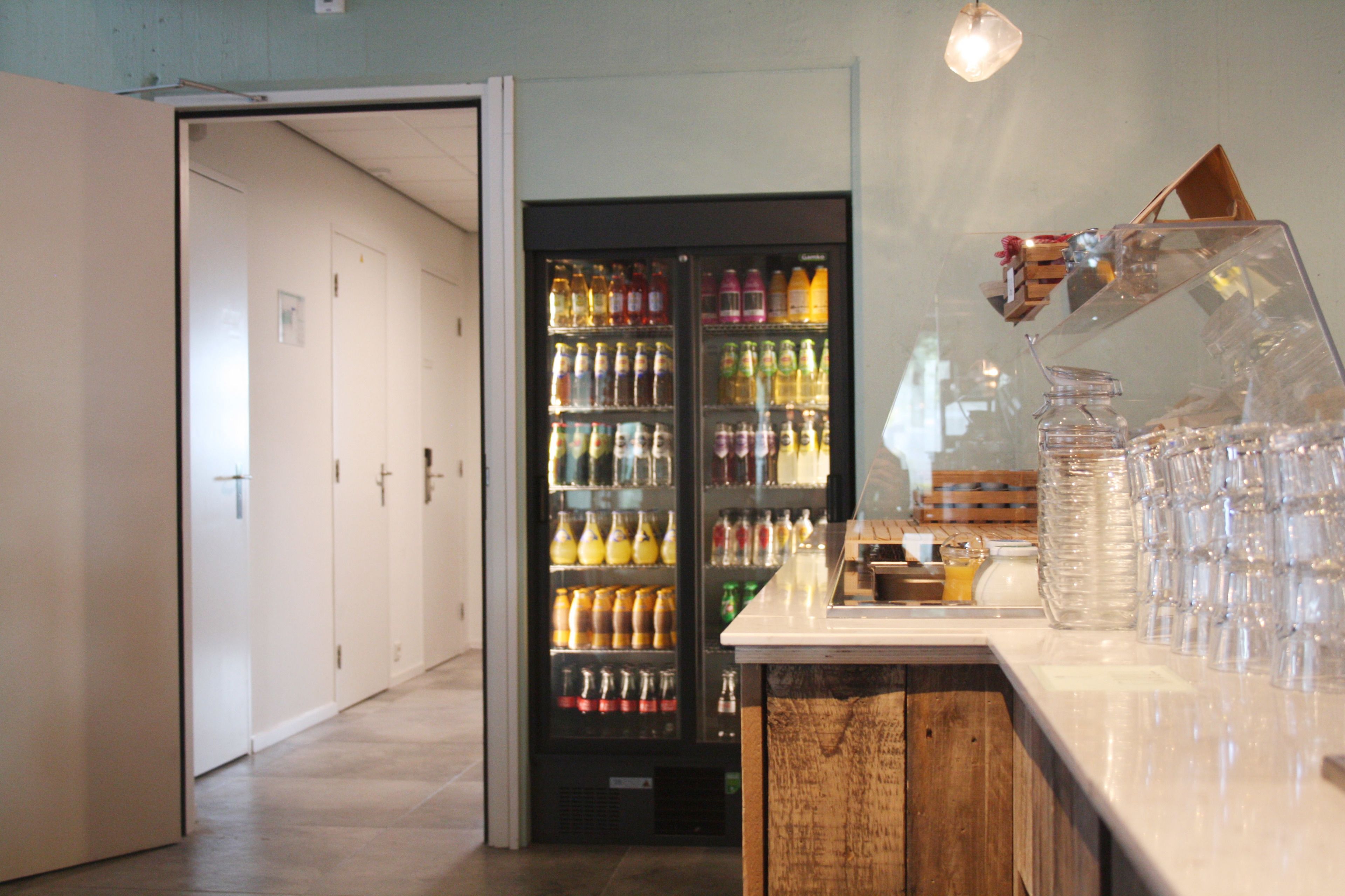 Un vestíbulo de hotel con un refrigerador lleno de bebidas, una barra de madera con cristalería y una puerta que da a un pasillo.