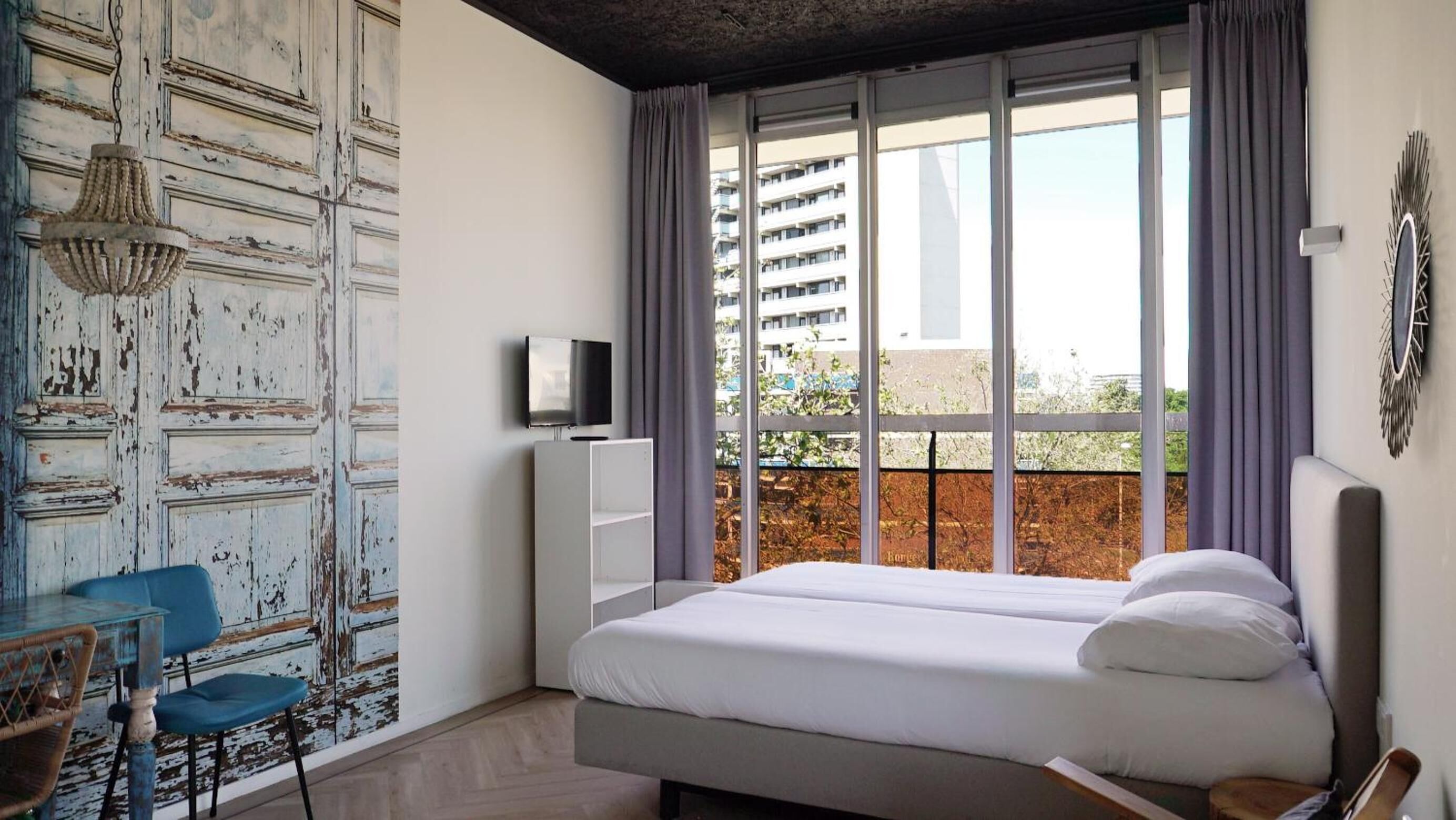 A bedroom with a bed, a TV, a desk with a chair, and a view of buildings through large windows.
