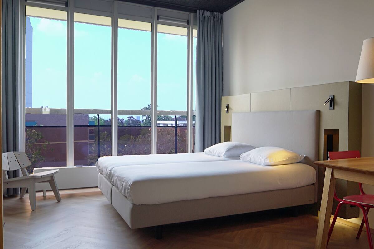 Een hotelkamer met een groot bed, een bureau, een stoel en een raam met uitzicht op gebouwen.