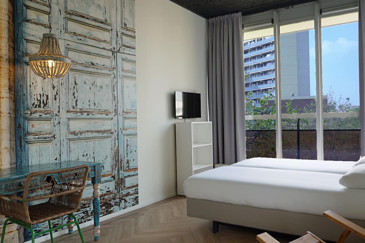 Een slaapkamer met een bed, een televisie, een raam met gordijnen, een stoel en een muur met een verweerde decoratie.