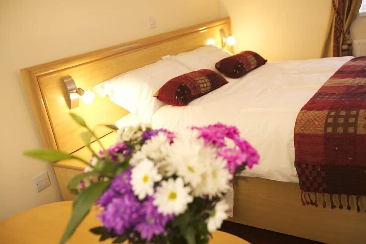 A neatly made bed with white and red pillows, a wooden headboard, and a bedside table with a vase of flowers.