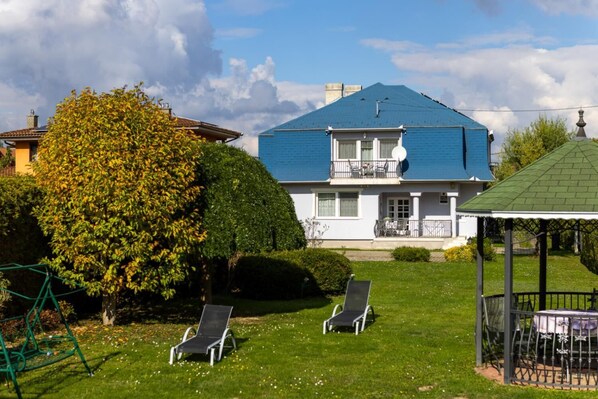 A two-story house with a blue roof, a balcony, and a small garden with two lawn chairs.