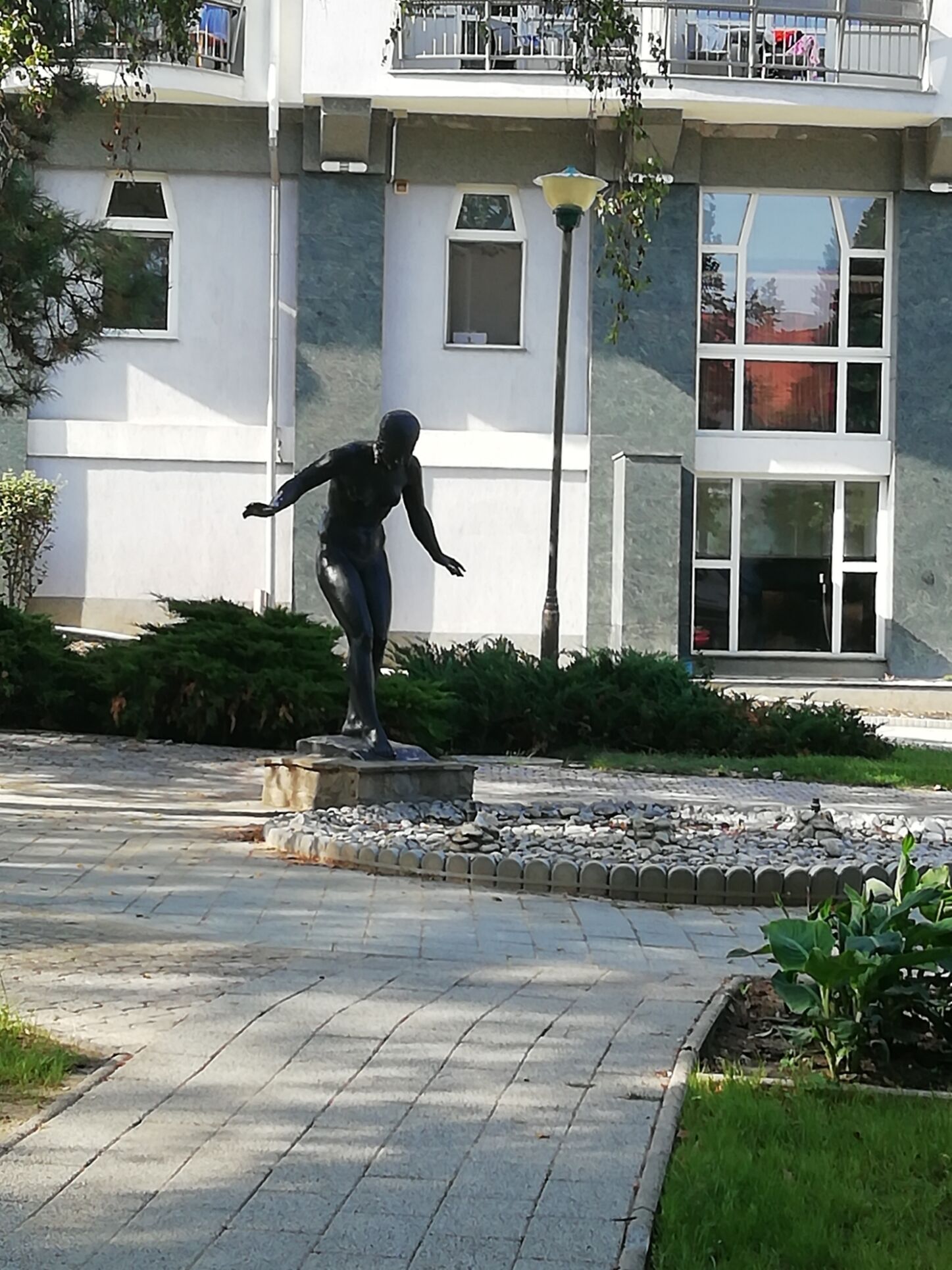 Una estatua de una persona en una pose dinámica sobre un pedestal en un área ajardinada, con un edificio al fondo.