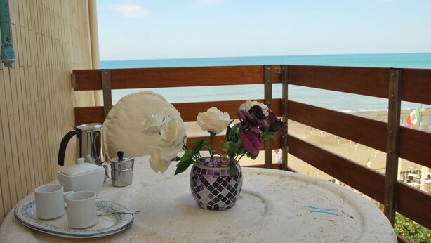 A balcony with a table set for tea, a vase with flowers, and a view of the beach. - Appartamento- Murex - Studio Apartment, Sleeps 3 (Marina di Castagneto Carducci)