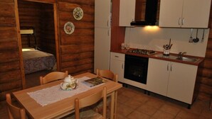 Interior acogedor de una cabaña de madera con una mesa de comedor preparada para dos personas, una cocina con armarios blancos y una estufa, y un pequeño rincón de descanso con una cama.