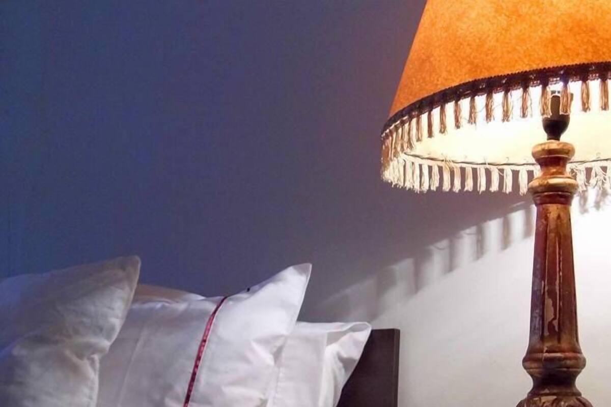 Una cama bien hecha con ropa de cama blanca y detalles rojos, una mesita de noche de madera con una lámpara y una pared oscura al fondo.