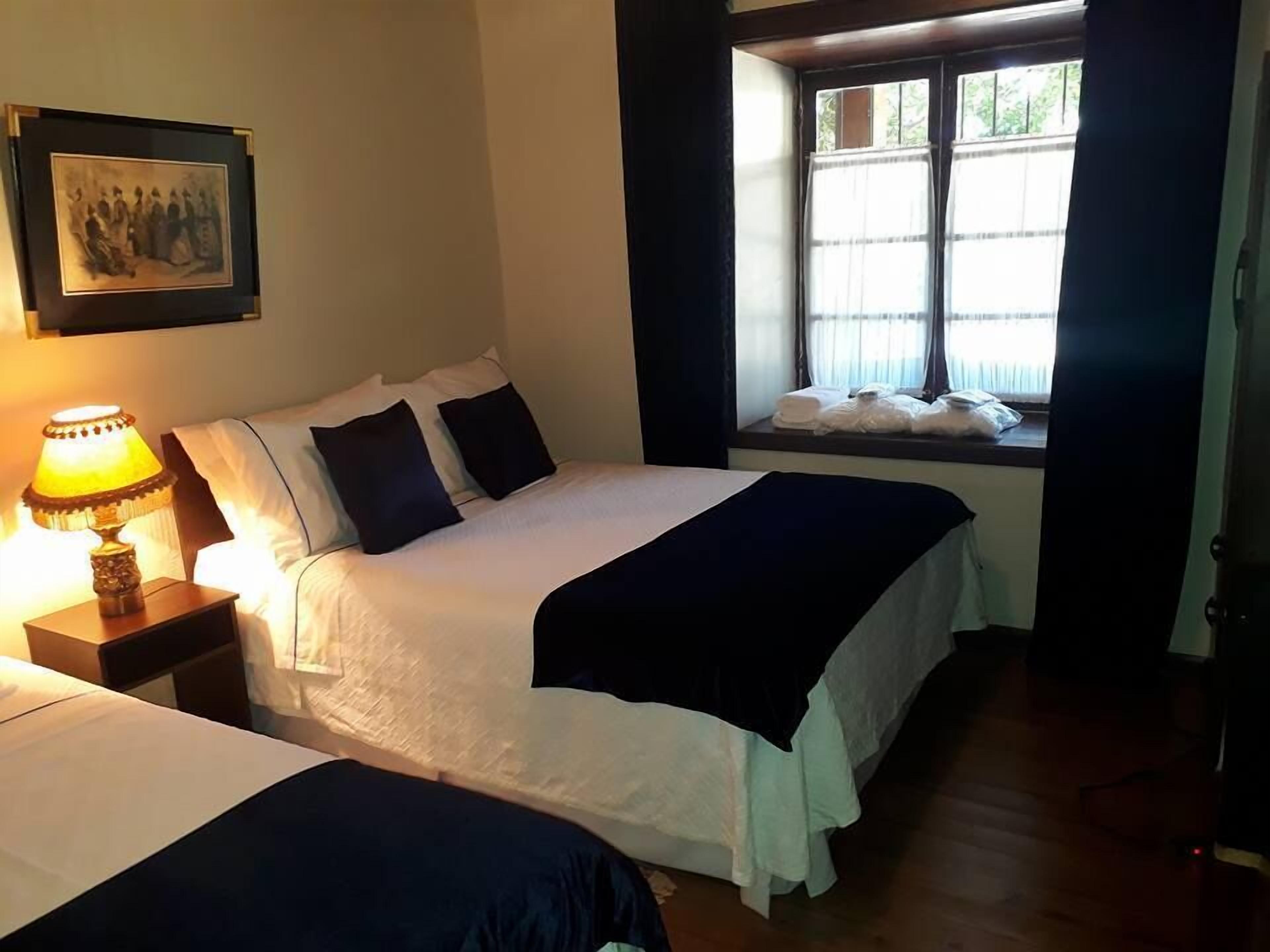 Habitación de hotel con dos camas, una mesita de noche con lámpara, una ventana con cortinas y un cuadro en la pared.