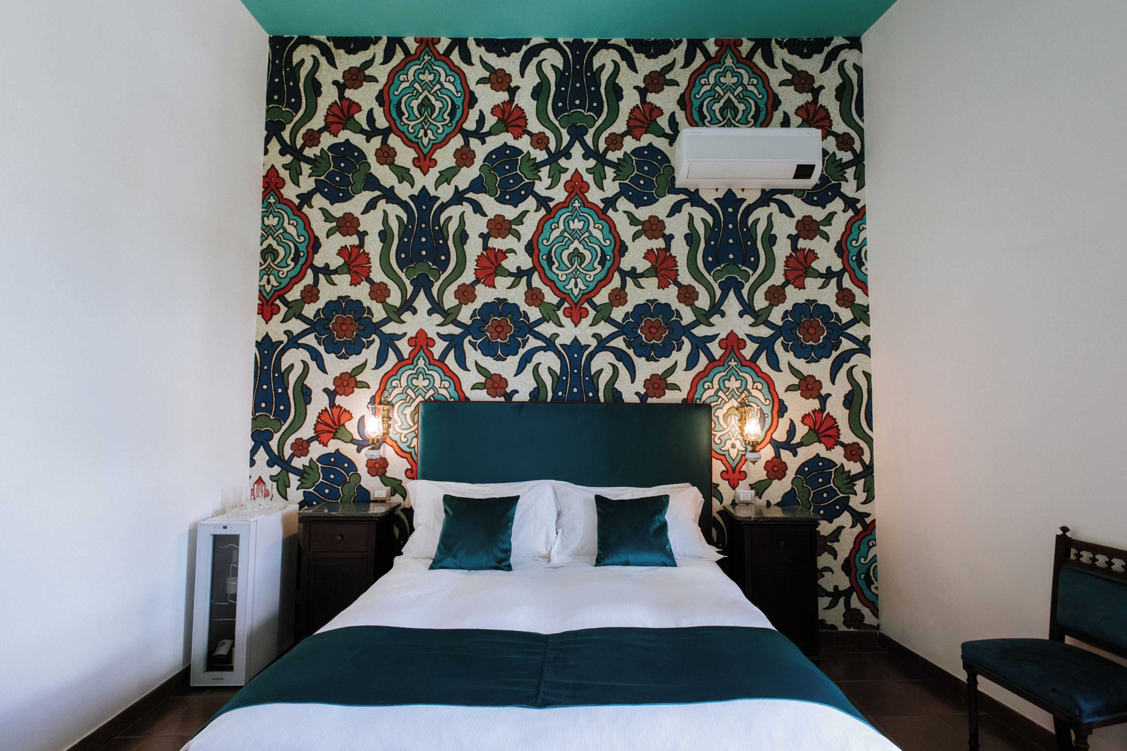 A neatly made bed with a green bedspread, a patterned wall, and a small bedside table with a lamp.