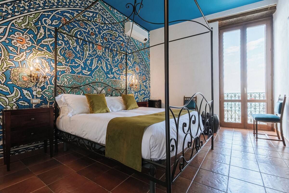 Un lit à baldaquin avec un couvre-lit vert, une table de chevet, une chaise et un mur orné d’un motif.