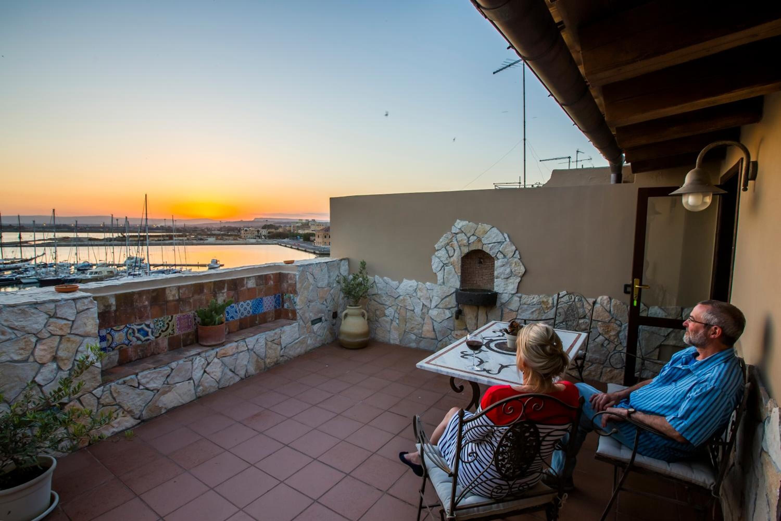 Eine Terrasse mit Steinmauern, einem Kachelboden, einem Tisch mit Stühlen und Blick auf den Hafen bei Sonnenuntergang.