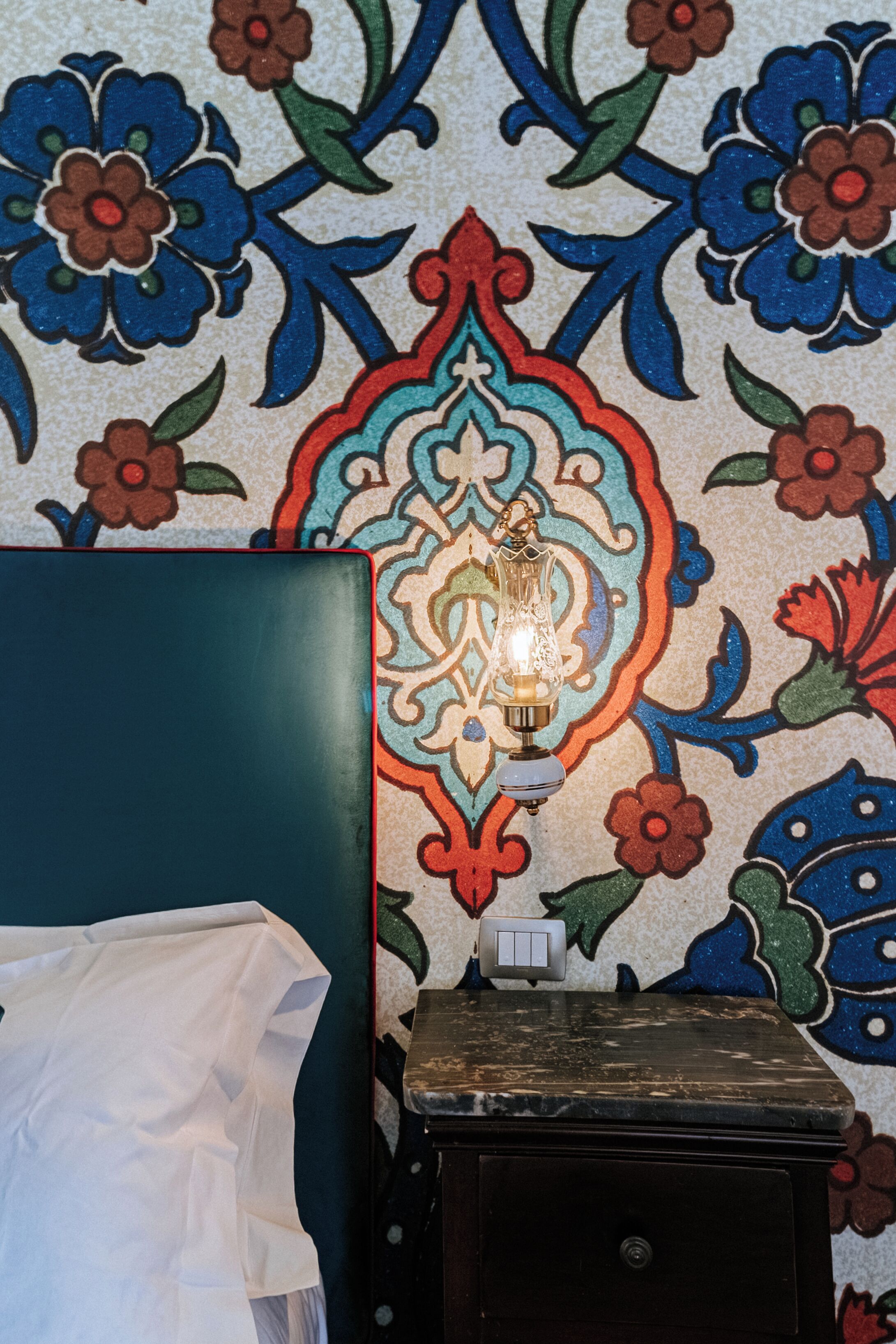 A room with a patterned wall, a bed with a white sheet, a small table with a light, and a switch.