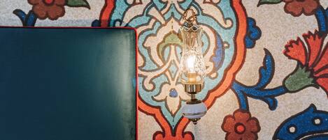 A room with a patterned wall, a bed with a white sheet, a small table with a light, and a switch.