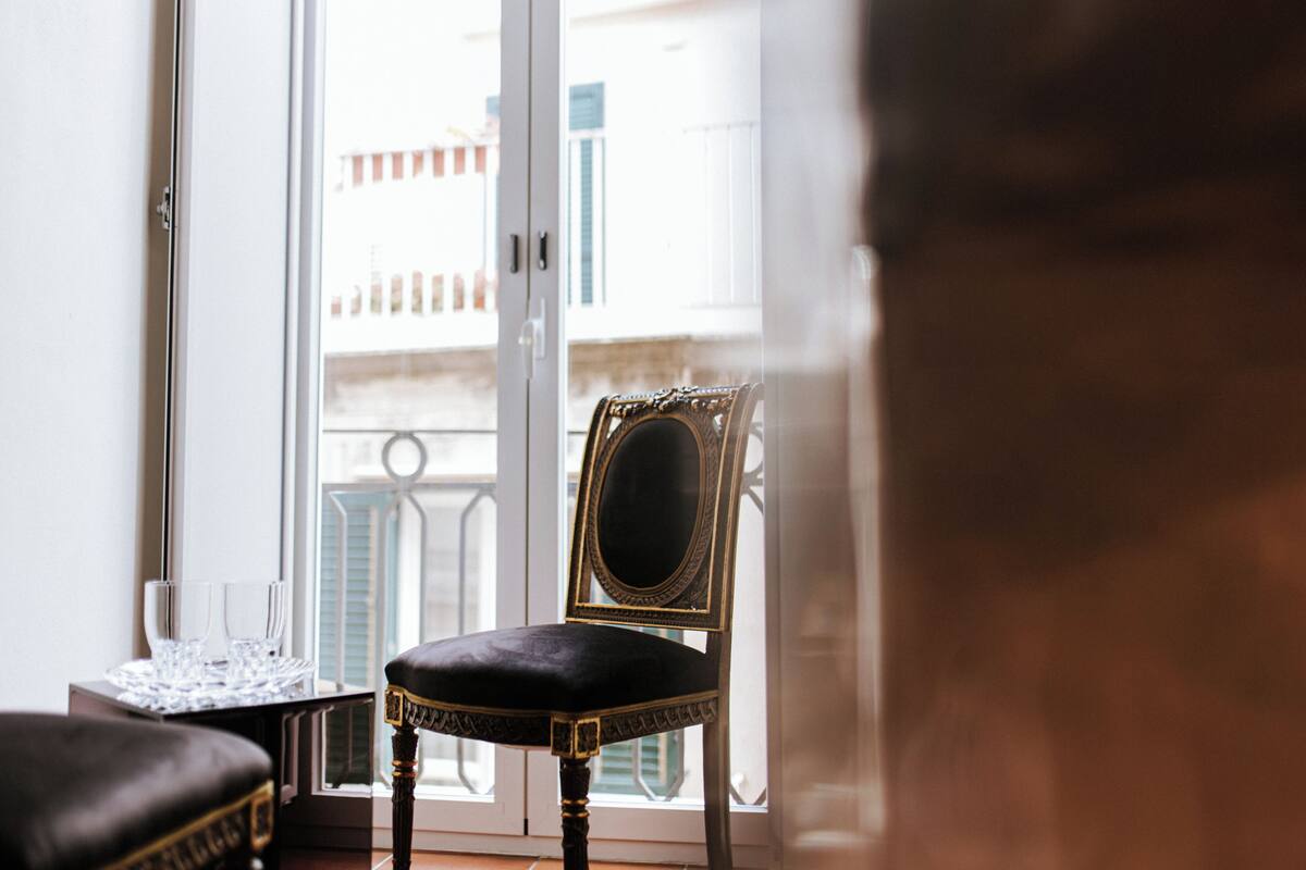Une chaise dotée d’un coussin sombre et d’un cadre orné, une petite table avec des verres, et une vue sur un balcon avec des bâtiments en arrière-plan.
