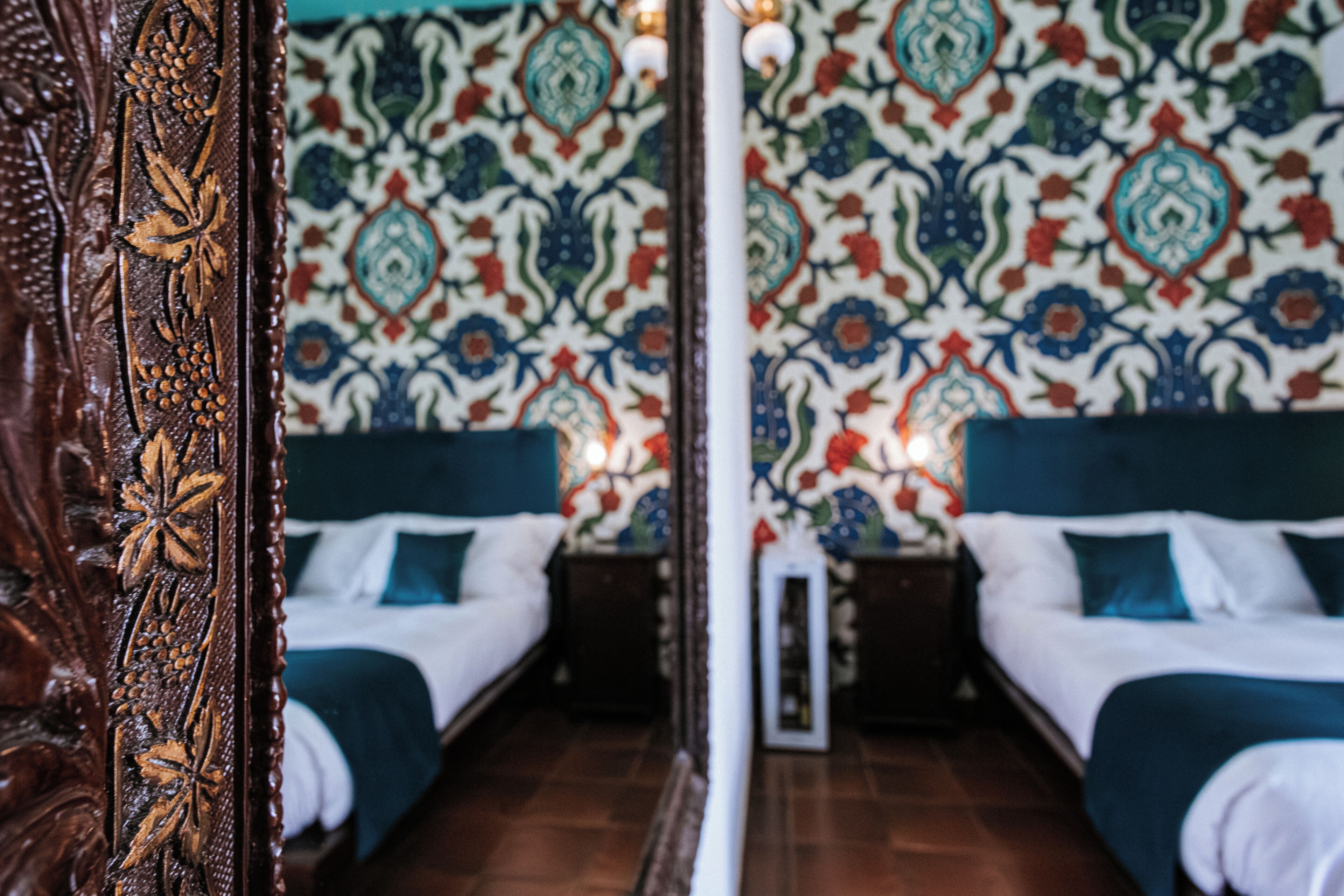 Habitación de hotel con pared espejada, dos camas con cabeceros azules y un papel tapiz estampado.