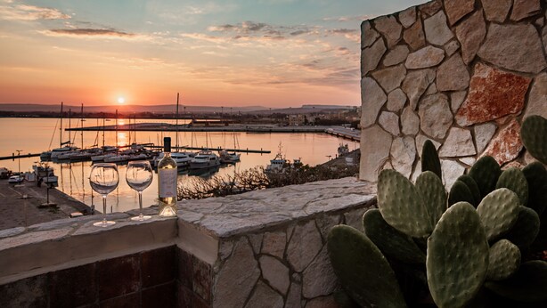 Een zonsondergang uitzicht vanaf een balkon met een stenen muur, cactussen en wijnglazen.