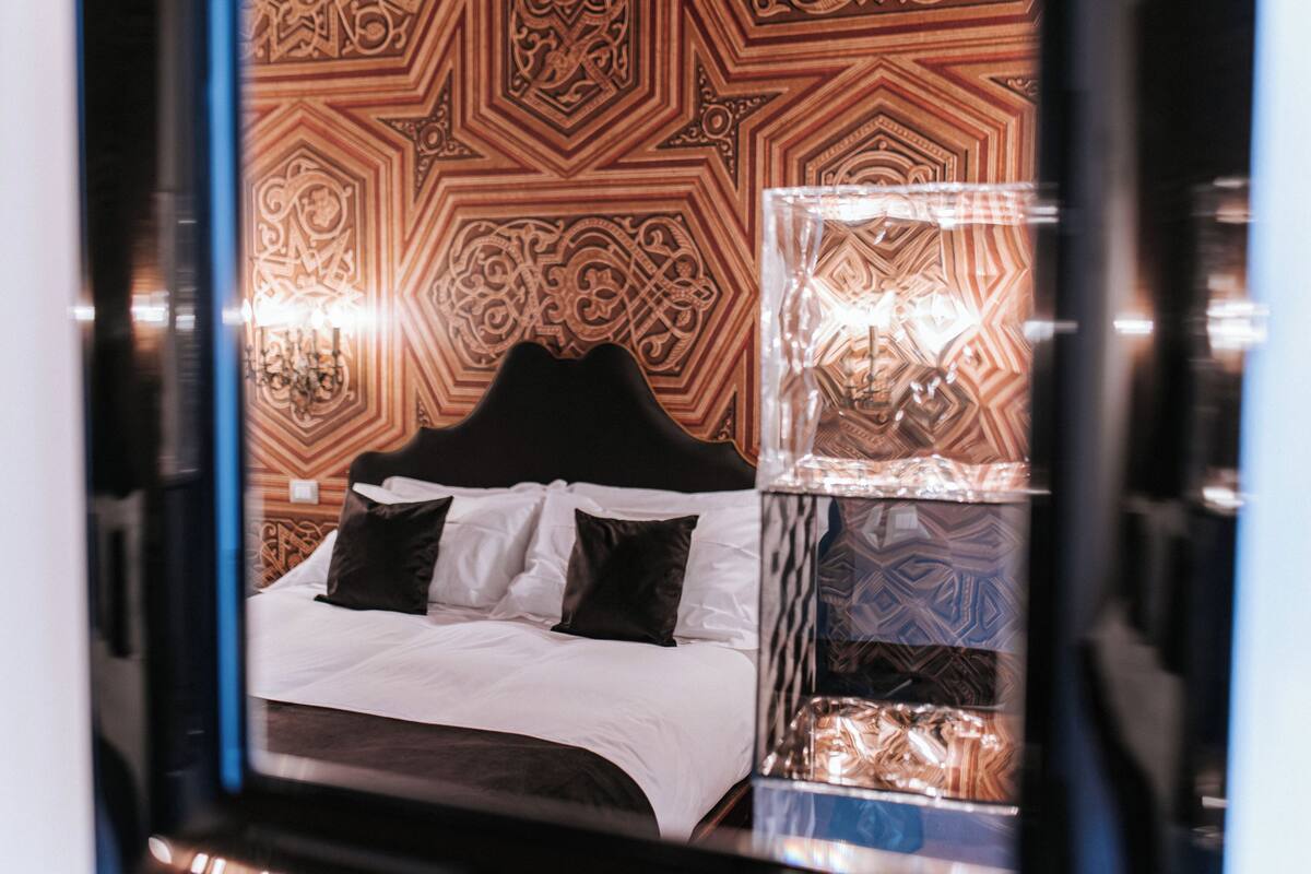 Un lit avec une literie blanche et des oreillers noirs, un mur à motifs et une table de chevet avec une lampe.