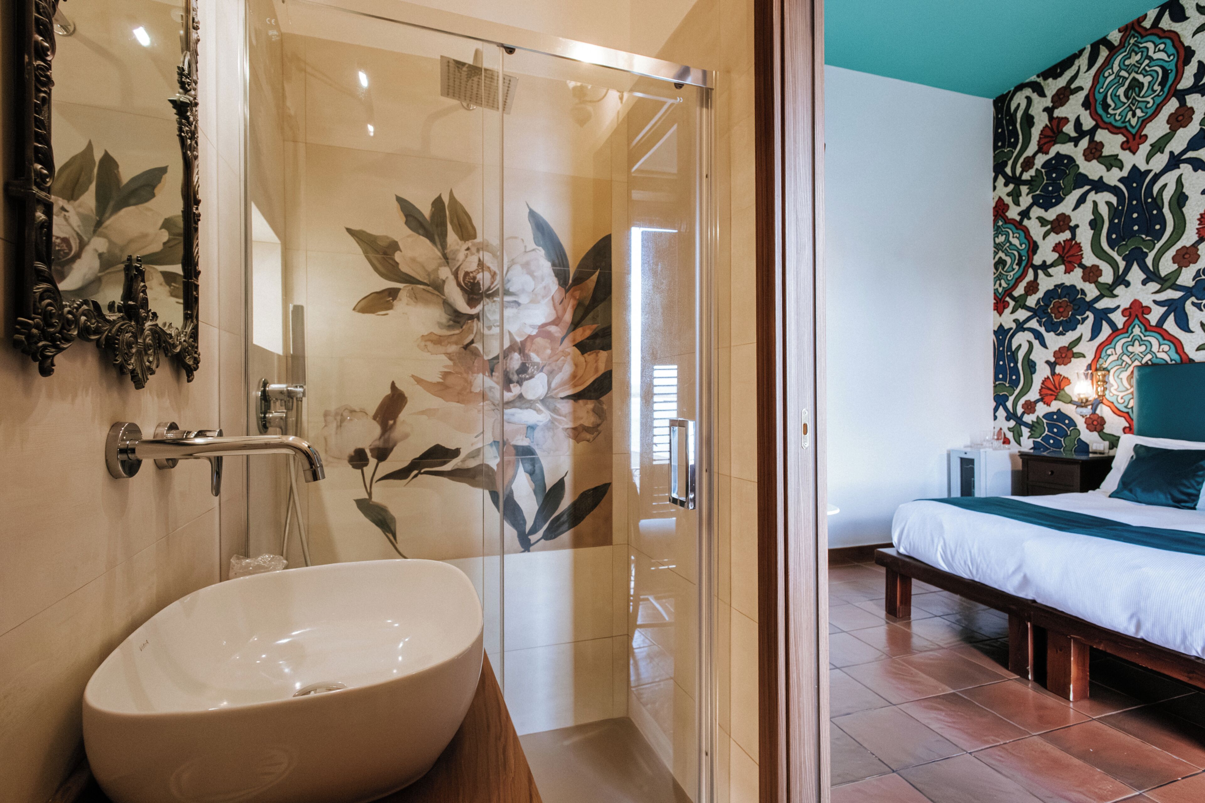 Ein Badezimmer mit einem großen, weißen Waschbecken, einer Duschkabine aus Glas und einem Bett mit blumigem Wanddekor.