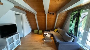 A modern loft bedroom with a sloped ceiling, wooden beams, a large window with curtains, a flat-screen TV, a sofa, and a small table. - Holiday park - Winnetou (Waldbrunn)