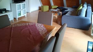 A cozy loft-style room with a wooden ceiling, a dining area with a table and chairs, a sofa, and a television on a cabinet. - Holiday park - Winnetou (Waldbrunn)