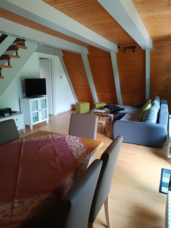 A cozy loft-style room with a wooden ceiling, a dining area with a table and chairs, a sofa, and a television on a cabinet. - Holiday park - Winnetou (Waldbrunn)