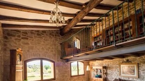A rustic interior with stone walls, wooden beams, a balcony with bookshelves, and large windows. - Hotel Hort De Fortunyo (Arnes)