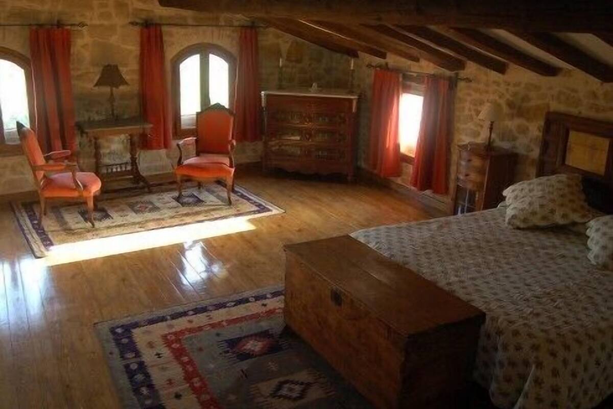 A rustic attic bedroom with exposed wooden beams, a stone wall, a wooden dresser, a bed with patterned bedding, and a red chair.