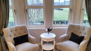 Two armchairs facing each other with a small table between them, a vase with flowers on the table, and a view of a green field through large windows. - Springfield Lodge B&B (Stirling)