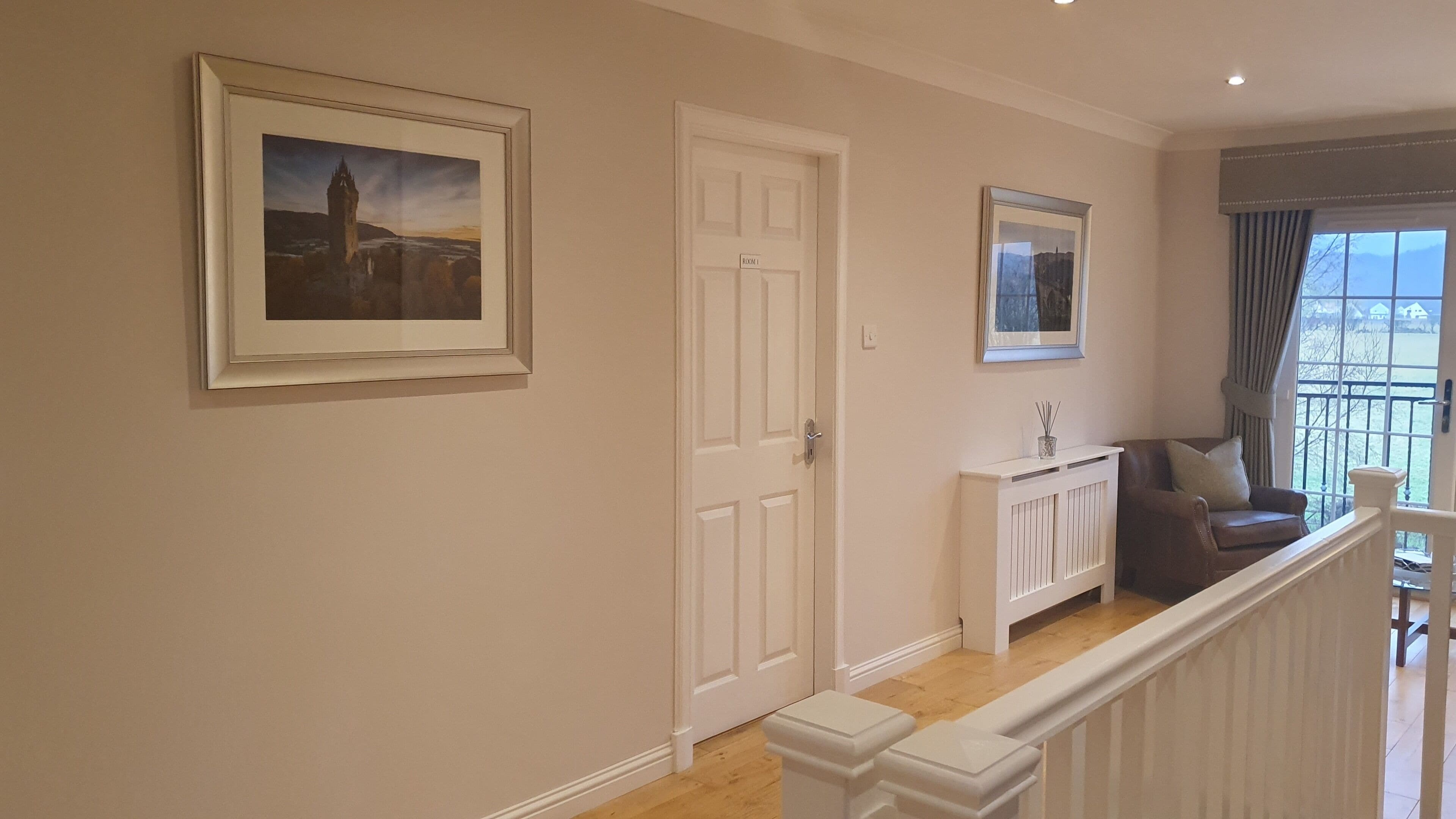 A hallway with a framed picture of a castle, a white door labeled "ROOM 1", a white console table with a vase, and a brown armchair by a window.