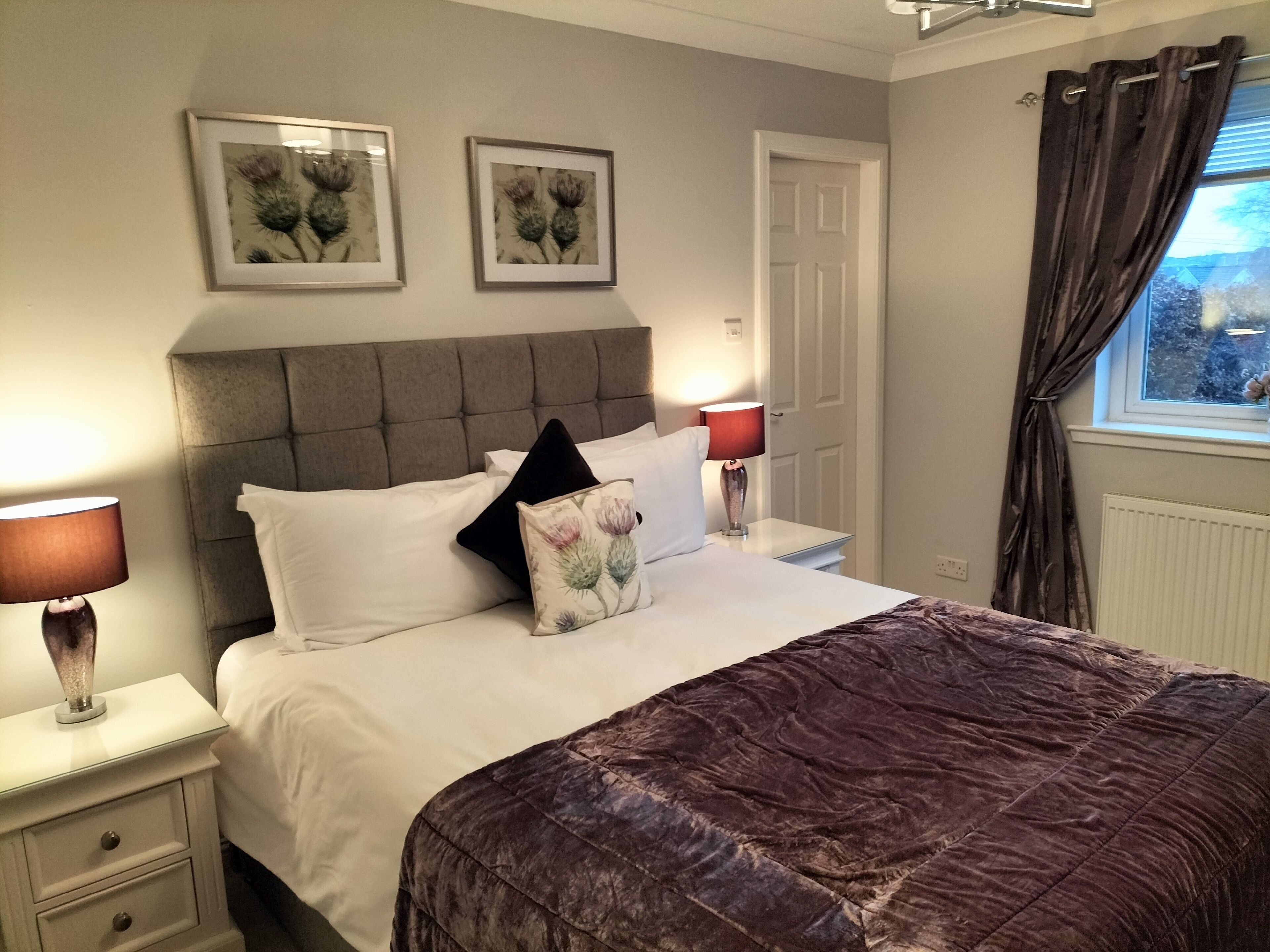 A neatly made bed with a dark brown quilt, two white pillows, and a decorative pillow with a floral design. There are two bedside tables with lamps, a window with curtains, and two framed pictures on the wall.