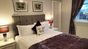 A neatly made bed with a dark brown quilt, two white pillows, and a decorative pillow with a floral design. There are two bedside tables with lamps, a window with curtains, and two framed pictures on the wall.