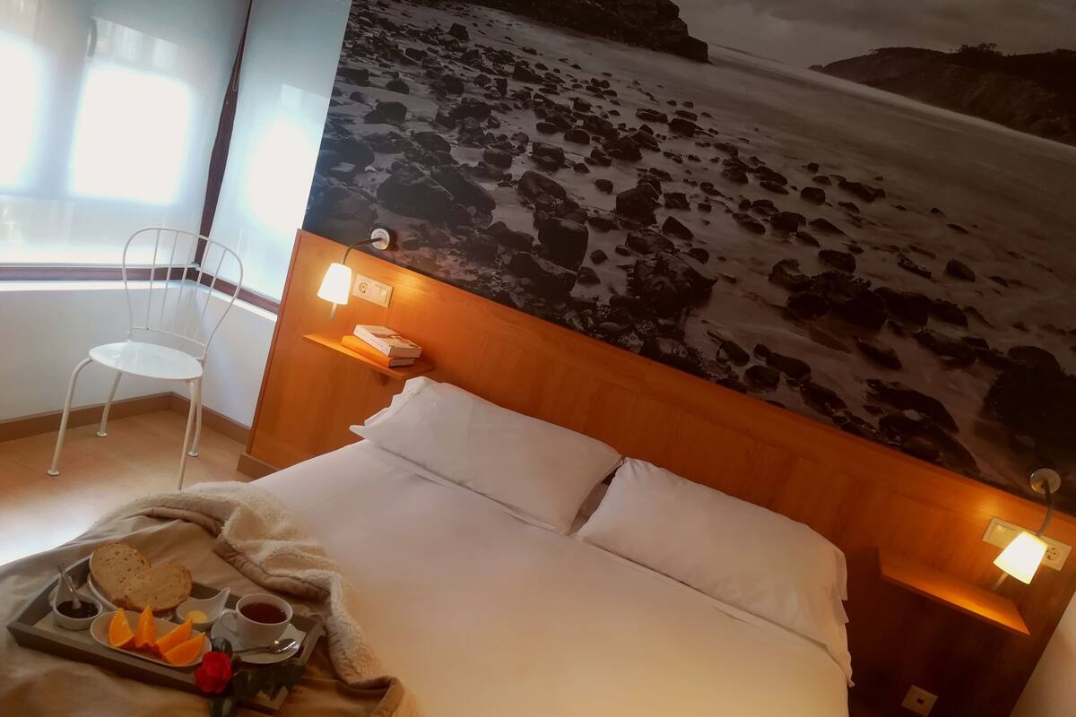 A hotel room with a bed, a tray with breakfast items, a chair, and a mural of a rocky beach.