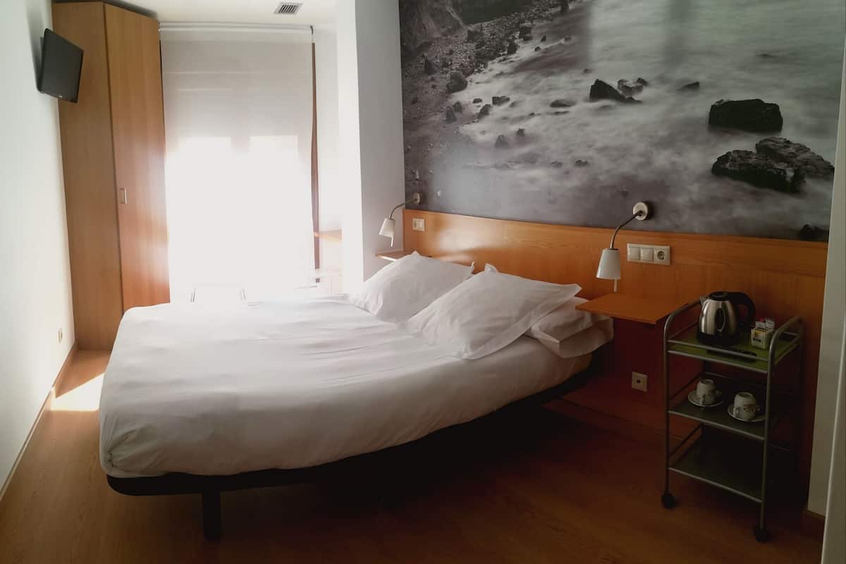 A neatly made bed with white linens, a wooden headboard, a wall-mounted TV, a bedside table with a lamp, a coffee maker, and a shelf with cups.