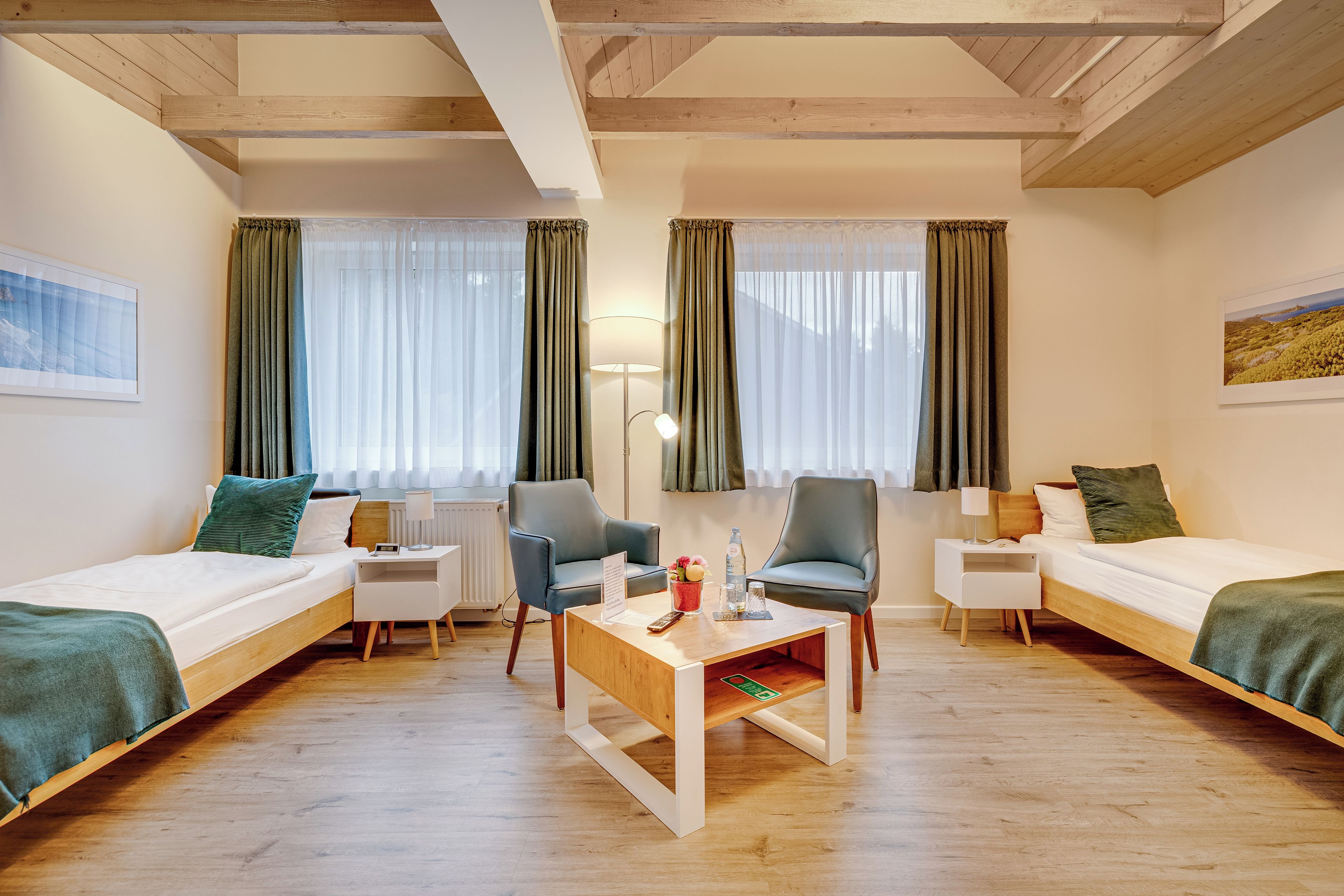 A hotel room with two single beds, a small table, two chairs, a nightstand, and a radiator.