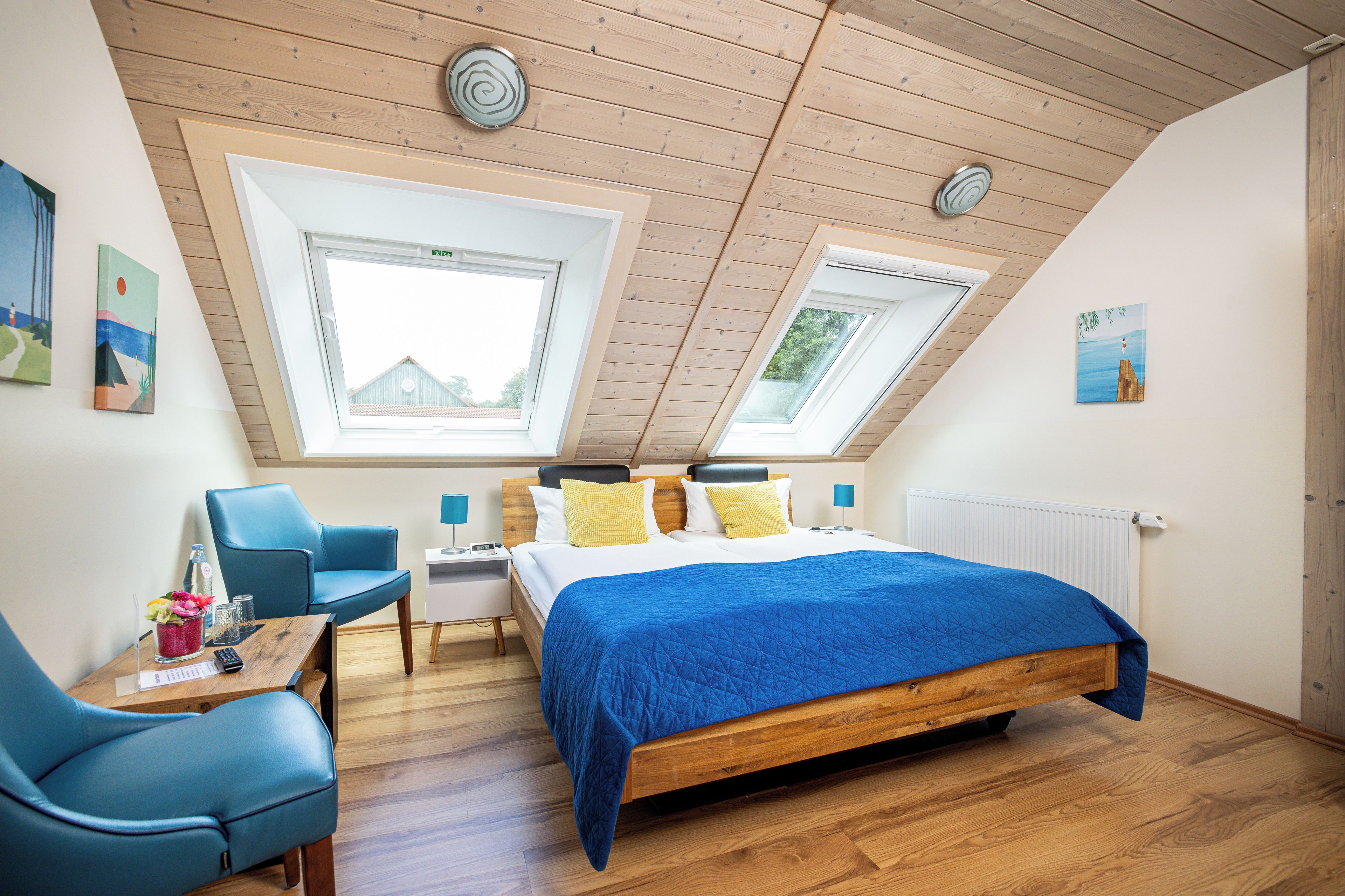 A bedroom with a sloped ceiling, skylights, a bed with a blue quilt, a desk with a chair, and artwork on the walls.