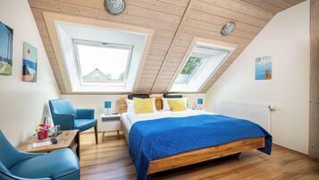 A bedroom with a sloped ceiling, skylights, a bed with a blue quilt, a desk with a chair, and artwork on the walls.