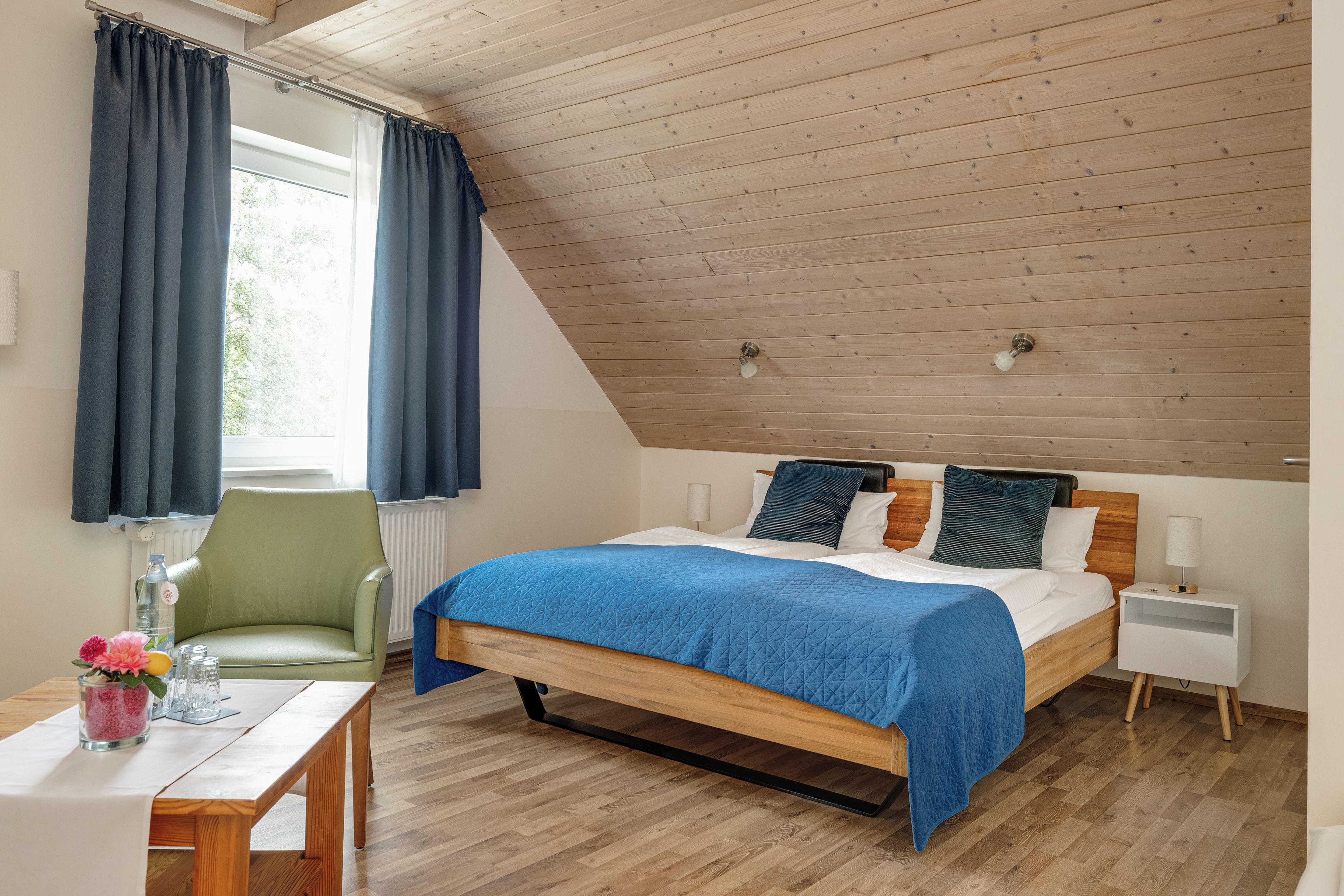 A bedroom with a sloped ceiling, a wooden bed frame, a bedside table, a chair, and a window with curtains.