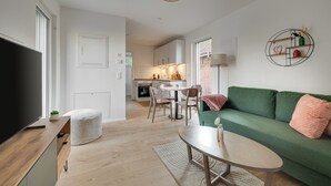 A modern living room with a green sofa, a coffee table, and a TV on a wooden stand. A kitchen is visible in the background with a dining area. - Haus Biederstaedt (Ottersberg)