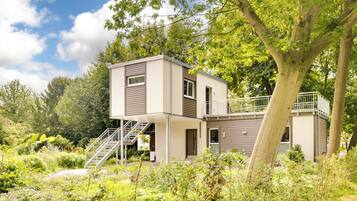 Une maison moderne à deux étages, dotée d’un balcon et entourée d’une végétation luxuriante et de plantes en fleurs.