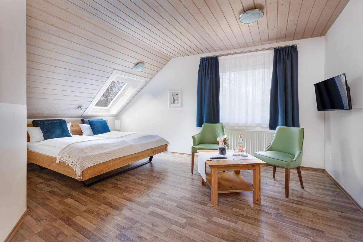 A bedroom with a wooden bed, a green chair, a wooden coffee table, and a TV on the wall.