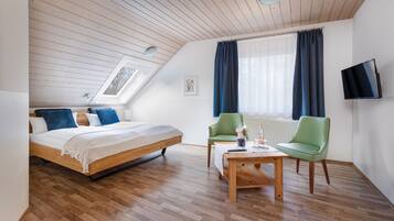 A bedroom with a wooden bed, a green chair, a wooden coffee table, and a TV on the wall.