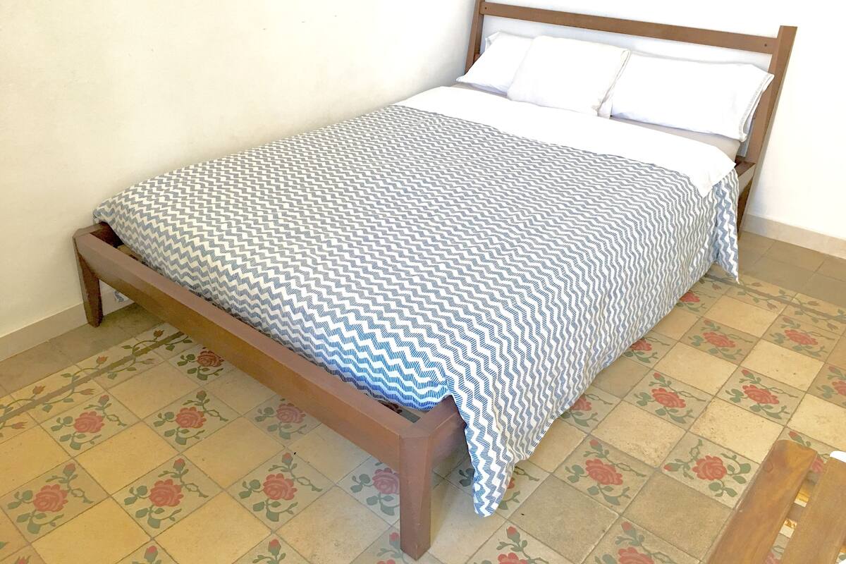 Uma cama com estrutura de madeira, roupa de cama branca e colcha estampada. O quarto possui pisos com ladrilhos estampados com motivos florais.