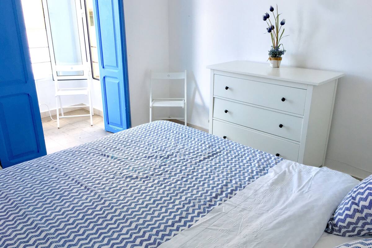 Quarto com uma cama, penteadeira branca, uma cadeira e um vaso com planta.