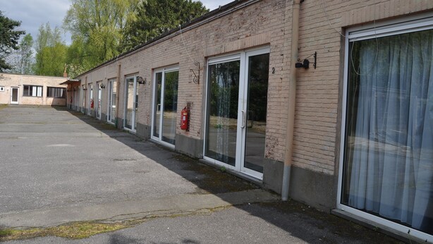 A row of single-story brick buildings with large glass doors and windows, a fire extinguisher mounted on the wall, and a clear sky in the background. - Logies Waasland (Sint-Niklaas)