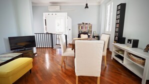 Una sala de estar con piso de madera, televisor, sofá, mesa de comedor con sillas, estantería y barandal blanco.