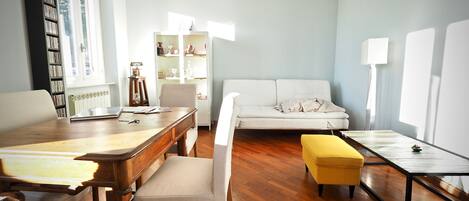 A living room with a wooden floor, a white sofa, a yellow ottoman, a wooden coffee table, a white armchair, a wooden desk with a laptop, and a bookshelf.