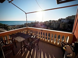 A balcony with a table and chairs, overlooking a coastal town with buildings and the sea.